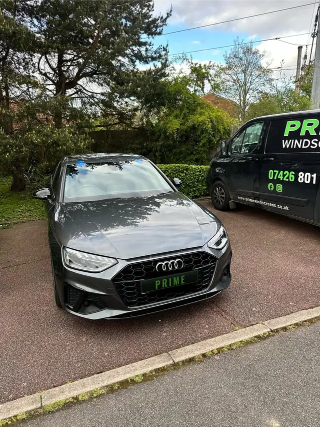Grey Audi A4 in a parking bay with blue tape on its new windscreen, the Prime Windscreens van parked alongside