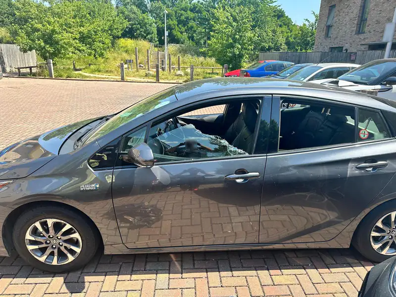 Grey Toyota Prius in a block paved car park with its front passenger window smashed and sagging in the frame