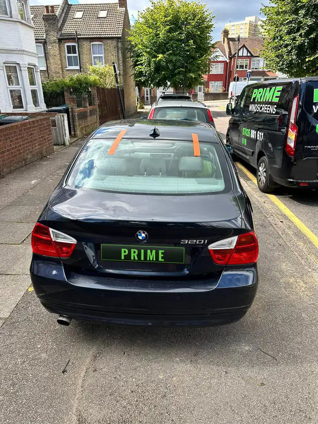 Dark blue BMW 3 Series saloon on a residential street with orange tape holding its new heated rear screen, the Prime Windscreens van alongside