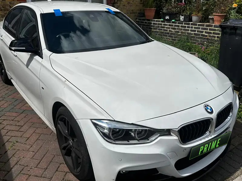 White BMW 3 Series on a block paved front drive with strips of blue tape holding its new windscreen while the bond sets