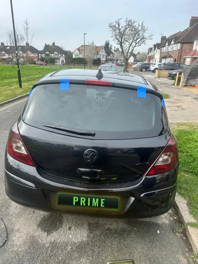 Black Vauxhall Corsa parked beside a grass green with blue tape holding its new rear screen